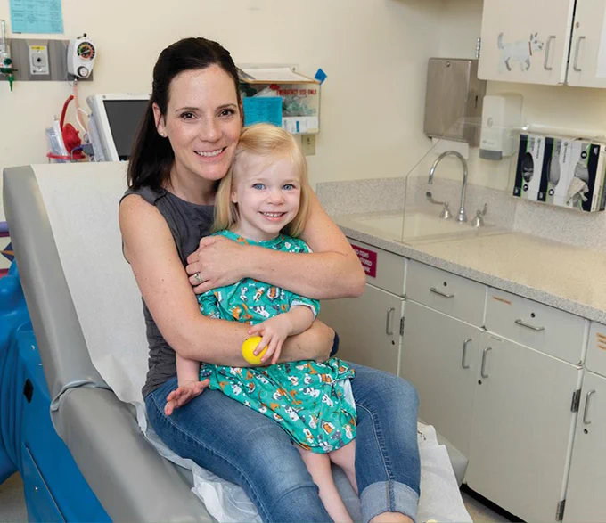Two people are smiling and sitting in a medical exam room. The adult is embracing a young child who is holding a toy. Both are casually dressed and look content. They are surrounded by medical equipment and cabinetry.