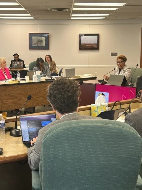Woman at a board desk speaking to a panel of others in the room