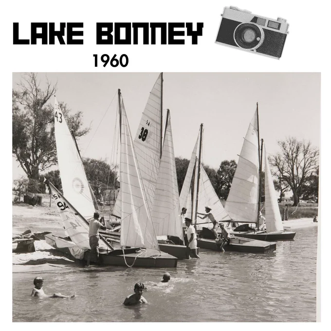 Image of sail boats lined up on the riverbank. Adults are stood in the water fixing sails on various ships. 
Children can be seen in the foreground swimming. 