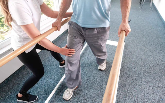 An allied health worker supports a man while he is walking along parallel bars in a hospital setting. Their heads are out of frame. 