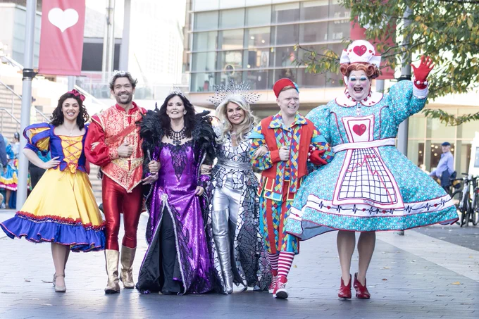Group of six performers in vibrant, eclectic costumes, including fairy tale and whimsical outfits, smiling and posing in a lively outdoor setting.