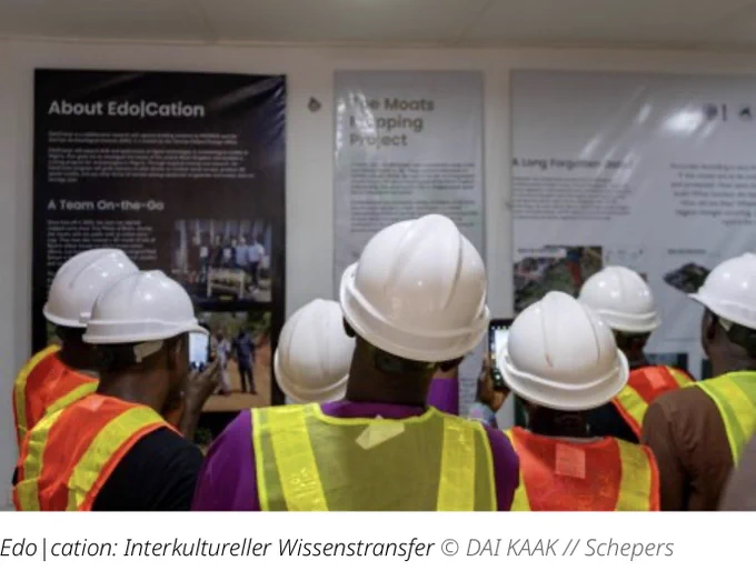 Foto einer Gruppe Menschen, von hinten gesehen, in Warnwesten und Schutzhelme gekleidet, die vor einer Reihe Informationstafeln zum Edo|Cation-Projekt stehen.