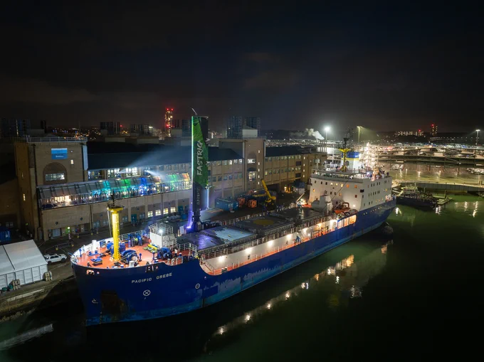 A large cargo ship docked at a brightly lit port at night. The ship features a distinctive, illuminated vertical structure resembling a sail. Behind the ship, there is a large building with illuminated windows, and city lights can be seen in the background.