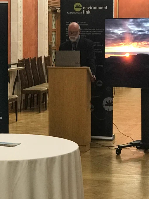 Photo of a man (Stephen Aston, chair of NI Environment Link) at a podium with a laptop, in Stormont, with a screen showing a beautiful sunset behind him, and an NIEL pop-up stand.
