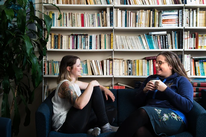 Two students facing one another on a couch in front of a bookshelf, drinking tea, smiling, and having a discussion.