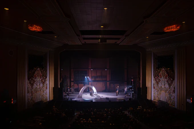 A wider shot of the theatre. The stage is surrounded by two large, rich, and ornamental art deco panels, one on either side, and a tall cavernous ceiling. The fully built, freestanding arch is on stage, light reflecting on the ice.