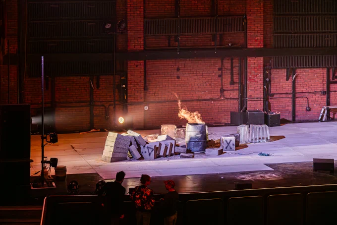 The remains of the arch on stage. Blocks of ice and concrete lie broken on the ground, as the fire underneath it swells. Three people silhouetted against the light stand watching and talking to each other.