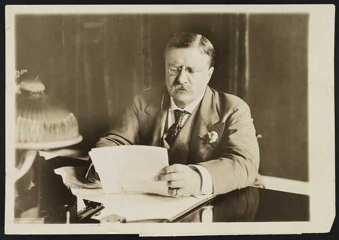 Theodore Roosevelt sits at a desk reading papers. He wears a mustache, glasses, and serious expression.