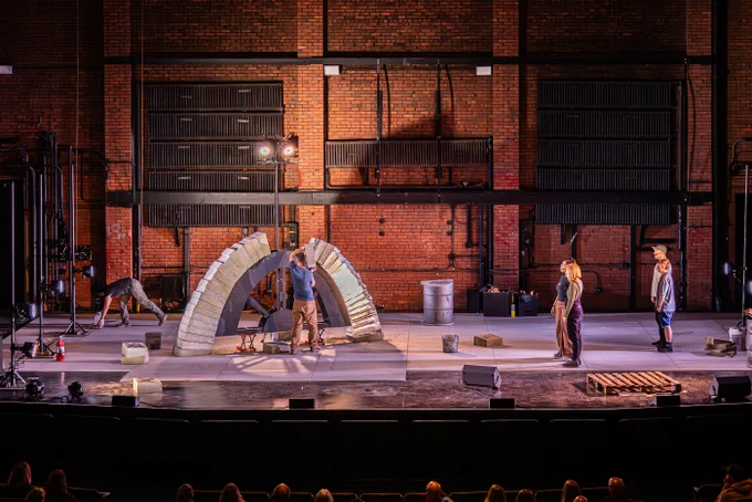 Indoors on a stage, Seth and Nao build an arch of concrete blocks and ice. To one side, four performers stand facing them, watching and singing. There is no scenery on stage, just the bare and industrial brick walls of the theatre. Lights are pointed at the arch, filling the space with shadows.