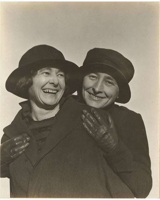 Portrait of Ida and Georgia O'Keeffe smiling in black hats and black coats, O'Keeffe stands behind Ida with her arms around her wearing black gloves.