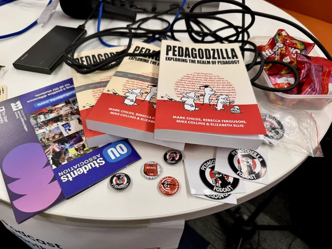 Table in the @Pedagodzilla podcast booth with some topical swag laid out (pins, books, and stickers).