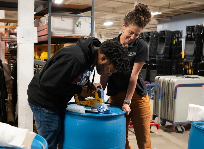 Two people work together to construct a rain barrel. One person uses a tool to cut a hole in the top of the barrel while the other holds the barrel still