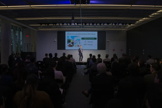 A person presenting on stage at a corporate event, addressing an audience seated in a dark room. The presentation slide shows graphs and text related to business strategies.