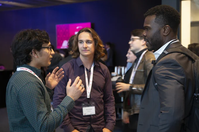 Three people engaged in a conversation at a professional event, wearing name tags and holding beverages.