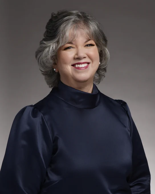 A smiling woman with chin-length grey hair wears a high-neck dark blue satin blouse. 