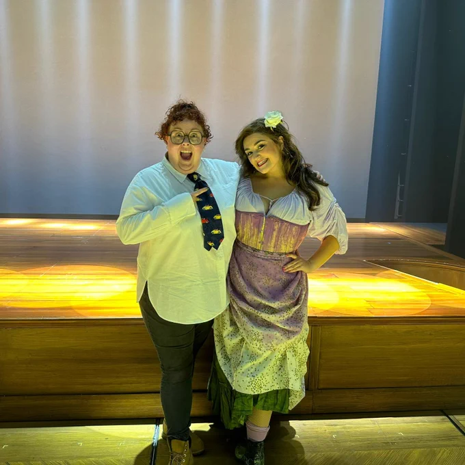 Lizzie and Daisy posing together on stage with bright lighting. Lizzie is wearing glasses, a white shirt with a tie, and dark pants, while Daisy is dressed in a colourful period costume with a flower in their hair.