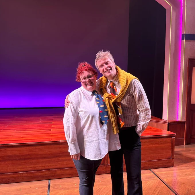 Lizzie and Stephen are posing, smiling, stand side by side on a stage with a purple background. Lizzie is wearing glasses, a white shirt with a tie, and dark pants, while the Stephen on the right, wears a yellow jumper around his neck as a scarf, a pinstripe shirt and a patterned tie.