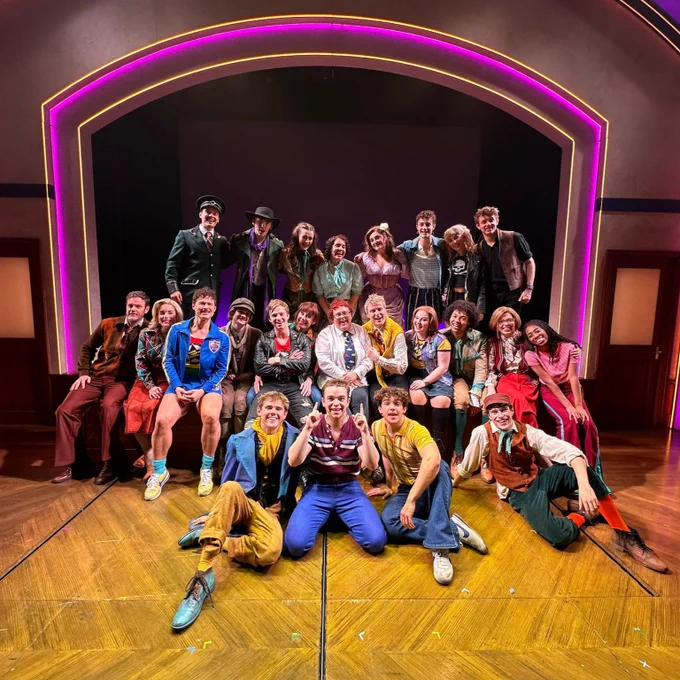 Group of actors posing on stage after a performance, smiling, with a brightly lit neon arch framing them in the background.