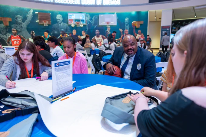 Secretary Tutwiler sits down with students at a table as they try and work out solutions to a challenge.