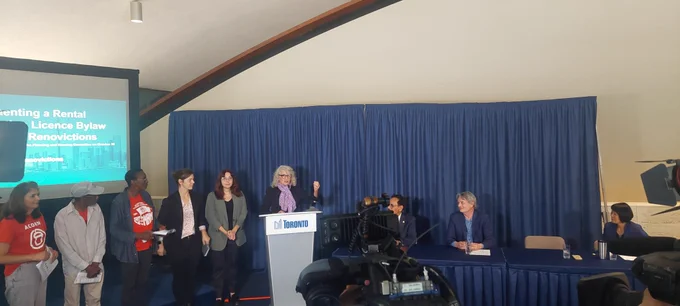 Councillor Fletcher at podium with five people standing beside her to her right and three people sat at table to her left speaking in front of news cameras.