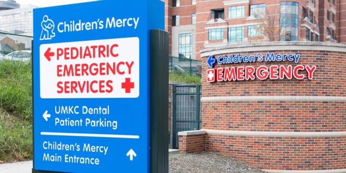 Directional sign at Children's Mercy, indicating locations for Pediatric Emergency, UMKC Dental Patient Parking, and the Main Entrance, with the hospital building in the background.