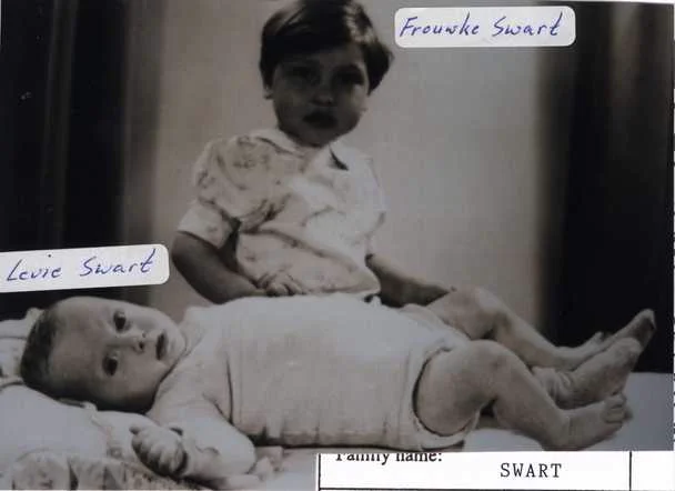 A young girl and her baby brother are on a bed. He is lying, she is sitting behind me. They are both looking in the camera. Their names: Frouwke and Levie Swart, are writtien in the photo.