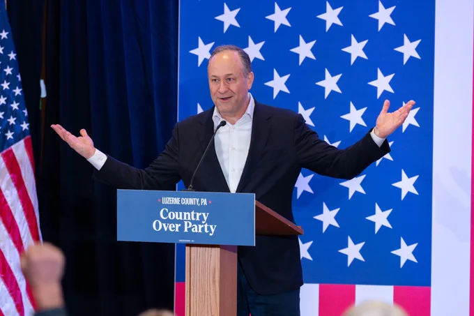 Second Gentleman Doug Emhoff speaking at a podium with a sign reading "Country Over Party" in Luzerne County, PA, with an American flag behind him. He is greeting the crowd with open arms and a smile on his face.