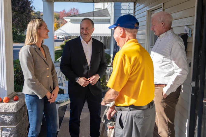 Second Gentleman Doug Emhoff chatting with supporters, smiling and conversing outside a their house on a sunny day. They are standing on the porch of the house.