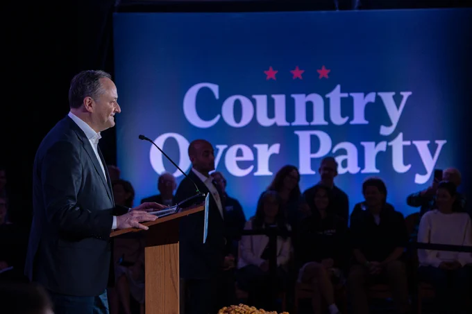 Second Gentleman Doug Emhoff at a podium during a "Country Over Party" event, with audience members seated in the background under a banner with the event's name.