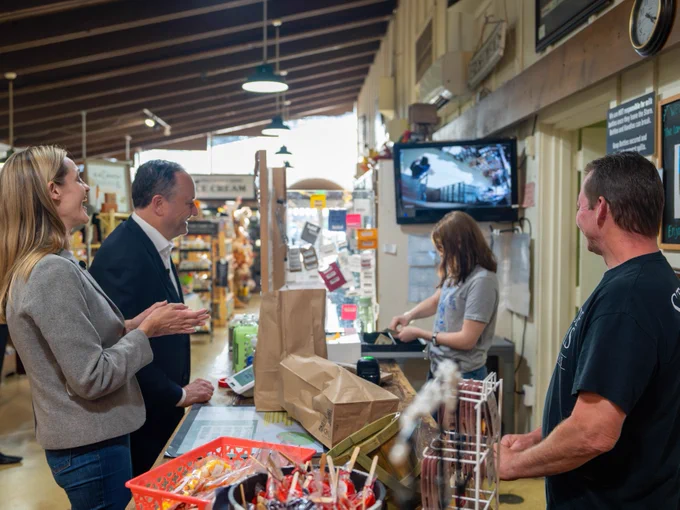Second Gentleman Doug Emhoff laughing with a cashier at a checkout counter in a local market.