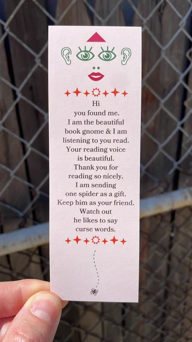 Bookmark featuring a strange little face at the top that’s wearing a red triangle hat. A tiny spider is at the bottom of the bookmark. Text reads:

Hi
you found me.
I am the beautiful book gnome & I am listening to you read. Your reading voice is beautiful. Thank you for reading so nicely. I am sending one spider as a gift. Keep him as your friend. Watch out
he likes to say curse words.