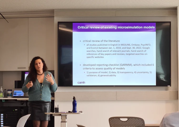 Dr Claire de Oliveira presenting to the left of a large screen. The screen shows a slide on "Critical review of existing microsimulation models".
