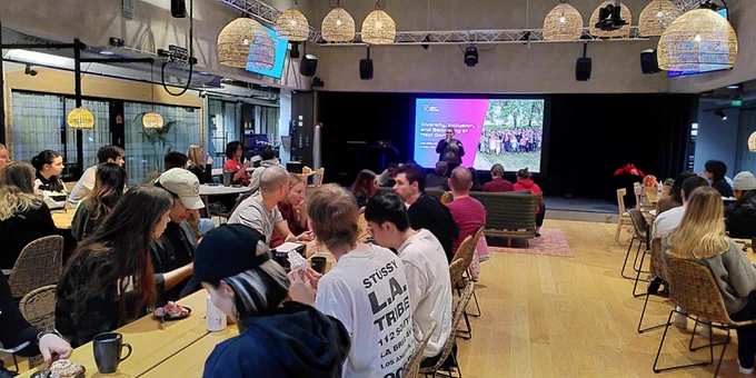 Students sitting in a cafe-like setting. Someone is on stage speaking into a microphone. 