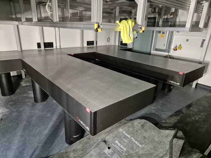 Laser optics table (chunky black table with a metallic top) sits below a set of metallic scaffolding - the gantry.