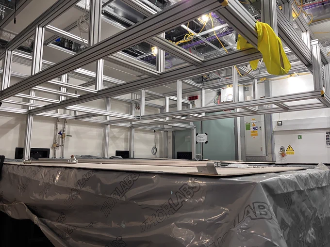 Laser optics table protected by plastic and card sits below a set of metallic scaffolding - the gantry.