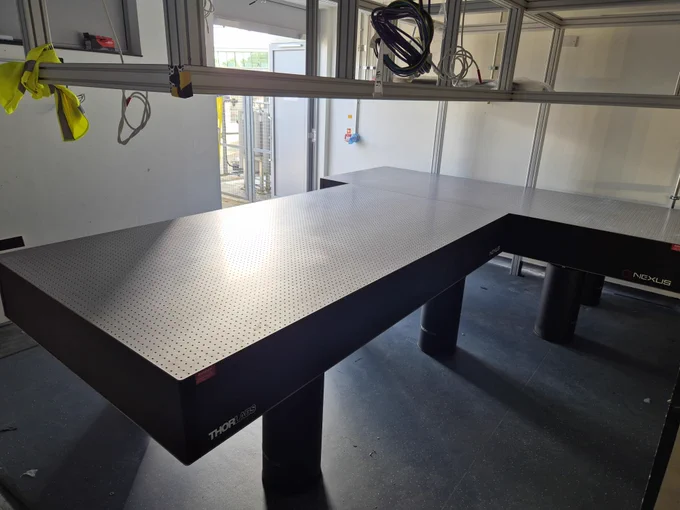 Laser optics table (chunky black table with a metallic top) sits below a set of metallic scaffolding - the gantry.