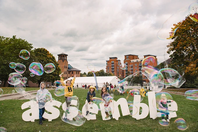 A group of children are playing with bubbles and jumping over a large sign that spells out Assemble. 