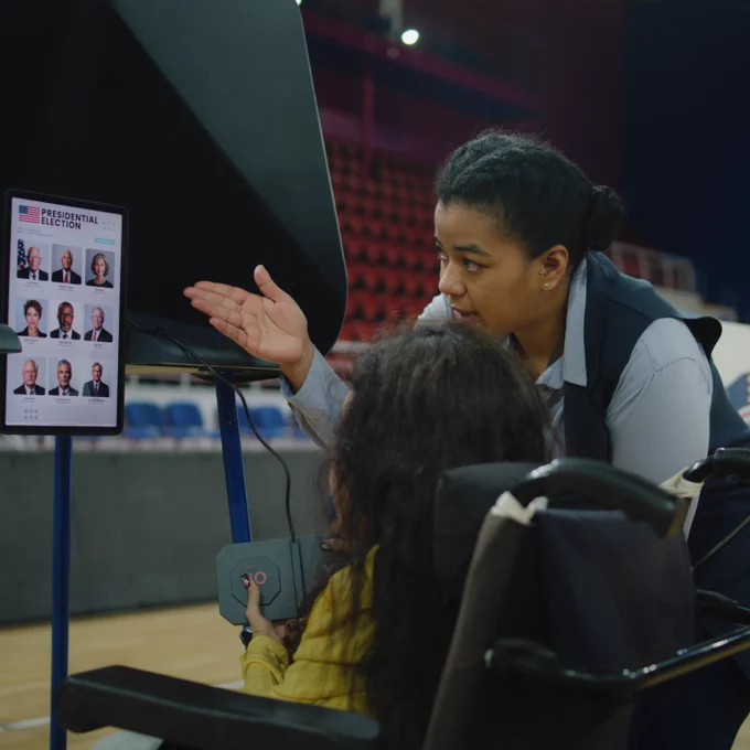 A poll worker, standing, gestures to a screen while a woman in a power wheelchair uses a device to vote at a polling place.
