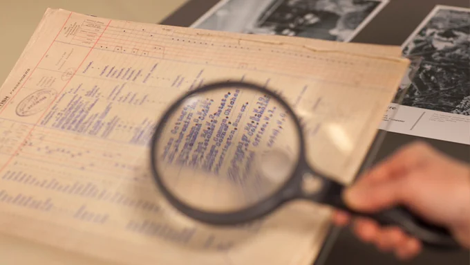 A hand holds a magnifying glass over some archival records, which are placed on a table, half covering some black and white photographs.

Photo courtesy of Migration Museum © Elzbieta Piekacz