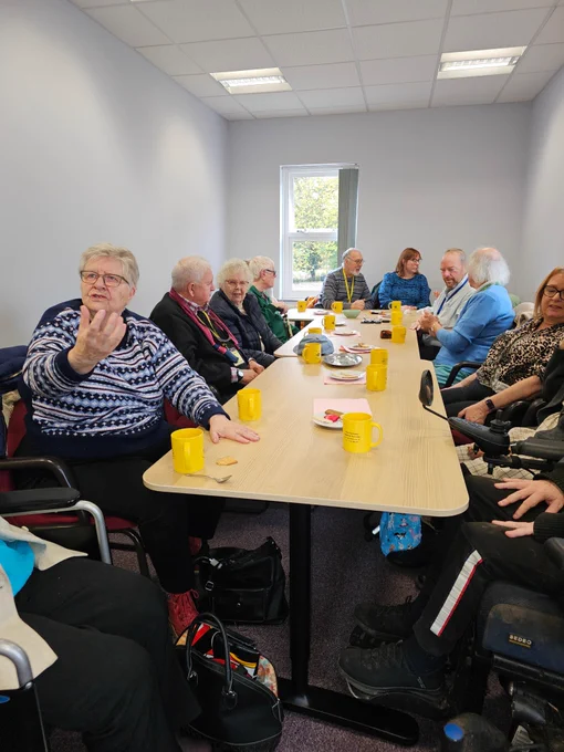A group of 11 people sat around a long table, some are in conversation and there are yellow mugs lined up and along the table where people are sat. There is also a selection of biscuits on plates lining the table as well.