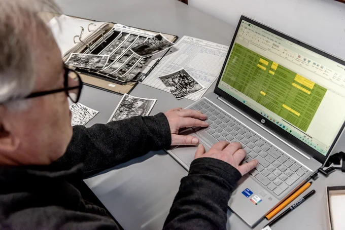 A person with glasses enters data on to a spreadsheet through a laptop. They are sat at a desk referring to black and white photographs and an open folder of papers.
