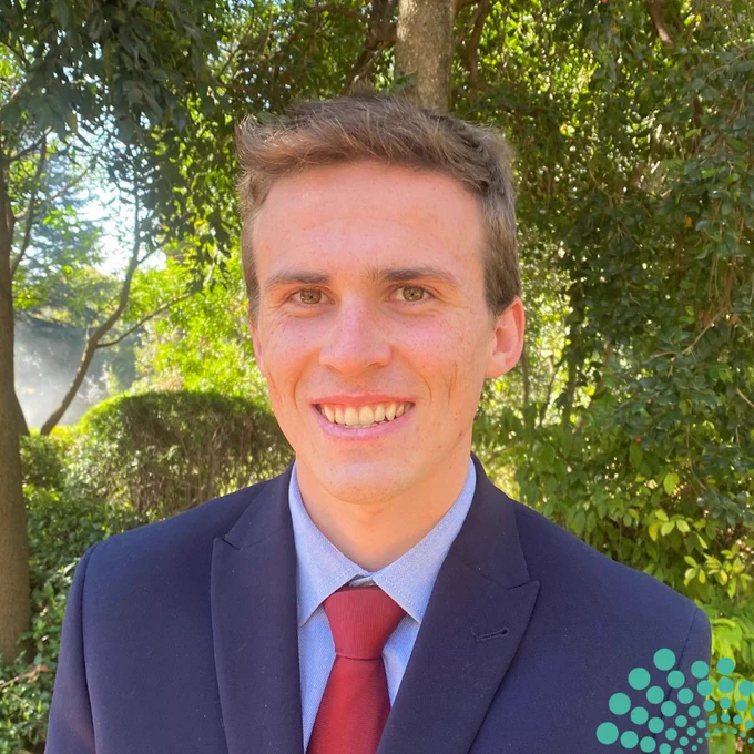 A young man is smiling warmly while standing outdoors in a lush, green setting. He is dressed professionally in a dark suit and red tie, exuding confidence and approachability. The natural sunlight filters through the trees in the background, enhancing the serene and vibrant atmosphere of the photo. The image captures both professionalism and a sense of connection with nature.