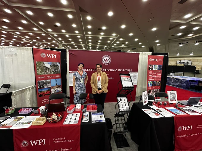 Two people standing in front of a large crimson backdrop advertising “Worcester Polytechnic Institute” with the school seal. There are smaller banners on either side advertising the school. There are tables in front of them with fliers and brochures.