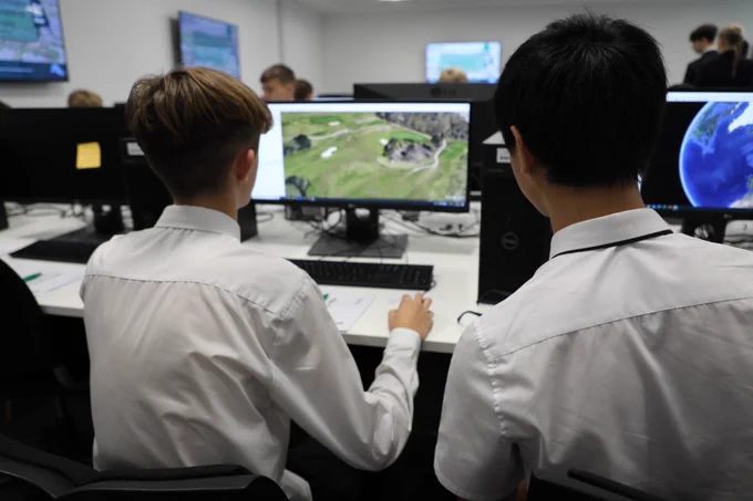 Two students in white shirts working at desktop computers in a classroom. Both are focused on a computer screen displaying a digital landscape. Other students and screens are visible in the background, indicating a classroom setting.