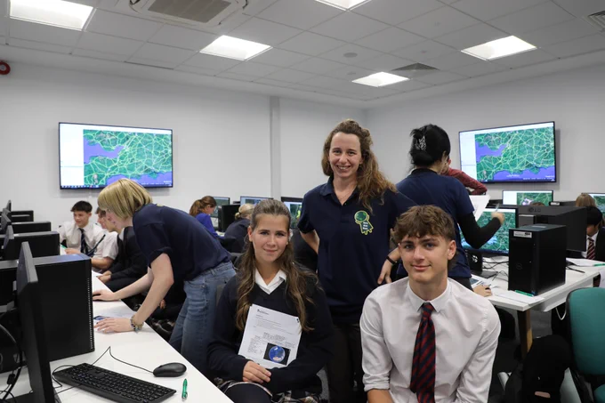 A classroom setting where students are working on computers. One student is holding a paper while seated next to another. An instructor is standing behind them, and computer monitors in the background display maps.