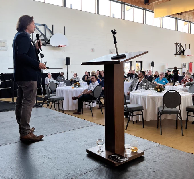 Dr. Peter Lewis speaking on stage with people sitting at tables in front of him