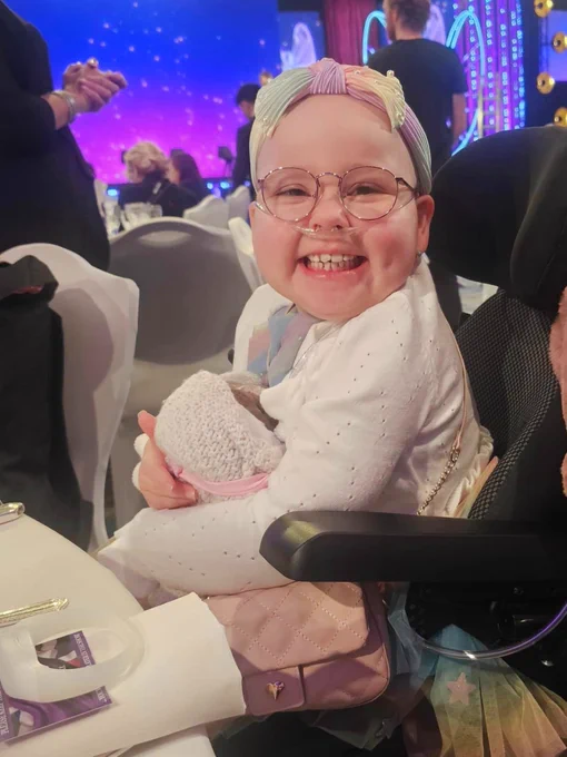 A young girl is sitting in a wheelchair wearing a bright coloured headband and beaming at the camera. 
