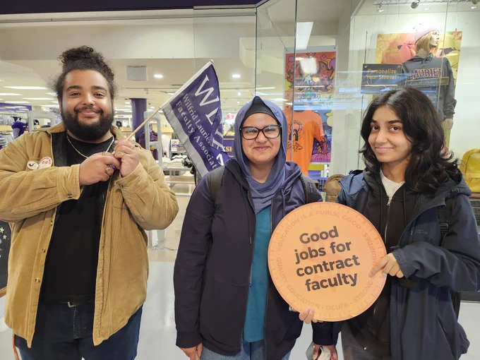 A photo of three students. One is holding a WLUFA flag, another is holding a sign that says "Good jobs for Contract Faculty"