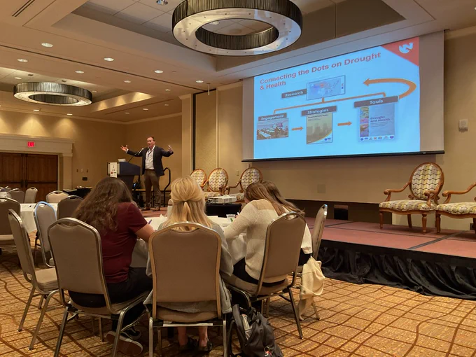 Dr. Jesse Bell stands on a stage speaking at the 2024 Nebraska Environmental Public Health Conference. Four people are pictured sitting at a table.