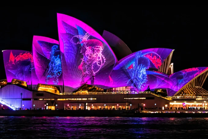 Sydney Opera House illuminated with vibrant purple lighting featuring abstract designs, viewed from across the water at night.
